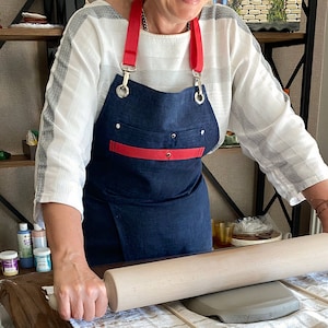 Op de afbeelding: Een persoon draagt een blauwe denimschort met rode bies en witte banden en rolt een stuk klei uit met een houten deegroller. De schort heeft een grote zak aan de voorkant. De persoon staat voor een houten tafel met verschillende knutselbenodigdheden.