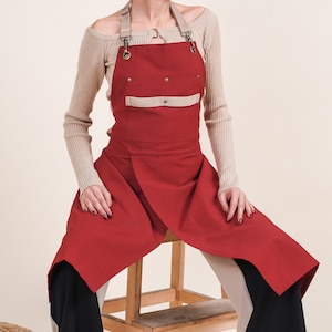 May include: A red apron with a beige pocket and adjustable straps. The apron is made from durable canvas material and has a relaxed fit.