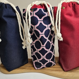Può includere: Tre sacchetti di stoffa con coulisse e manici in corda bianca. I sacchetti sono blu scuro, blu scuro con un motivo geometrico bianco e rosa, e rosso.