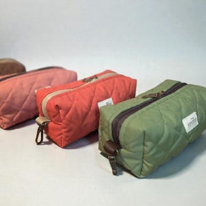 May include: A collection of quilted pencil cases in various colors, including pink, coral, and olive green. Each case features a diamond pattern, a zipper closure, and a small fabric tag. The cases are designed for storing pens, pencils, or other small items.