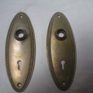 May include: Two oval-shaped brass door plates featuring a beaded border design. Each plate includes a central circular opening for a door knob, a keyhole opening below, and two small screw holes at the top and bottom for mounting purposes.