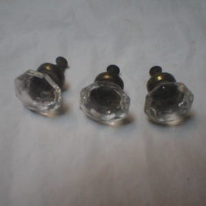May include: Three clear glass knobs with an octagonal shape and a faceted surface. Each knob features a dark metallic base with a central screw attachment, providing a functional hardware component for cabinets, drawers, or furniture pieces.