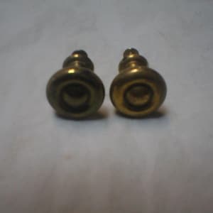May include: Two matching brass cabinet knobs with a circular, rounded face and a tiered, stepped base. The metal has a warm, aged gold-toned finish with a smooth, polished surface texture. Each knob features a central raised circular detail.
