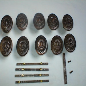 May include: A set of ten oval-shaped, ornate, bronze-colored door knobs with a decorative floral design. The knobs are arranged in two rows of five. There are also four metal rods with small brass screws on each end.