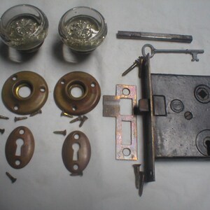 May include: A set of door hardware including two clear glass knobs with a starburst pattern, two circular brass rosettes, two oval keyhole escutcheons, a rectangular metal lock box, a metal key, a square spindle, and several small mounting screws.