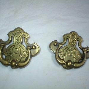 May include: Two antique brass drawer pulls with ornate floral designs. The pulls have a ring handle and are in excellent condition.