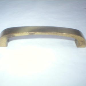May include: A curved, brass-toned cabinet pull handle. The handle has a rounded, arched design with two rectangular mounting posts. The finish has a slightly aged appearance, suggesting a vintage or antique style. Ideal for furniture restoration or adding a classic touch.
