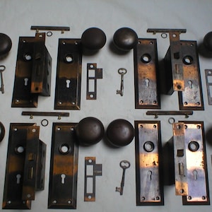 May include: Collection of antique door hardware, including bronze-colored doorknobs, rectangular faceplates, mortise locks, keys, and strike plates. The hardware has a vintage aesthetic, with a mix of dark and lighter bronze tones.