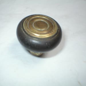 May include: A dark brown, round door knob with a brass-colored top. The top has concentric circles and a small, circular center. The knob is likely made of wood and metal, and is a classic design.