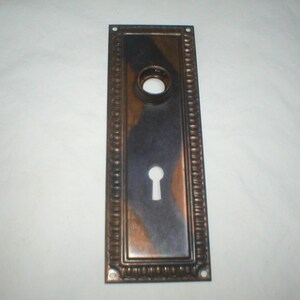 May include: Rectangular antique door plate with a keyhole and a circular opening. The plate has a dark, metallic finish with copper and bronze tones. The edges are decorated with a beaded pattern.
