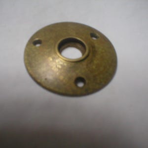 May include: A circular, antique brass-toned metal flange featuring a slightly domed profile. The component includes a central circular opening and three smaller, evenly spaced mounting holes positioned around the perimeter of the disc.