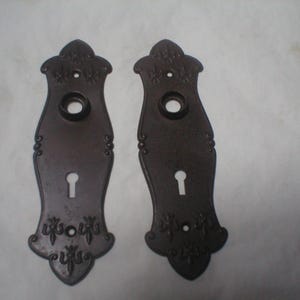 May include: Two dark brown metal door plates with an elongated, curved shape. Each plate features a circular opening at the top for a handle, a keyhole below, and decorative embossed fleur-de-lis patterns at the top and bottom ends.