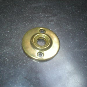 May include: A circular brass door rosette with a polished metallic finish. The hardware features a central opening for a spindle and two smaller, vertically aligned screw holes at the top and bottom for mounting to a door surface.