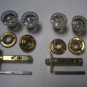 May include: Two sets of door hardware, each including a clear, faceted glass knob, a circular brass-toned metal rosette, a tubular latch mechanism, and a square metal spindle. The components are arranged in two matching groups on a plain surface.
