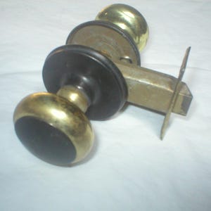 May include: A door knob set featuring two spherical brass knobs with black circular centers, connected by a central metal latch mechanism. The hardware includes a rectangular faceplate and circular rosettes, showcasing a combination of polished brass and matte black finishes.