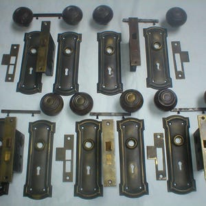 May include: An assortment of antique door hardware, including doorknobs, backplates, and latches. The hardware is made of metal with a dark bronze finish and a decorative design. The collection includes various sizes and styles of door hardware.