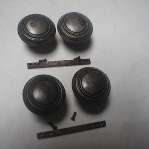 May include: Four circular dark metal door knobs with a beaded edge detail, accompanied by two rectangular metal mounting plates and three small screws. The hardware has a weathered, aged finish with visible signs of patina and surface wear.