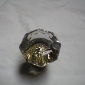 May include: A clear glass doorknob with a faceted design. The knob has a slightly yellowed tint and a raised, textured center.