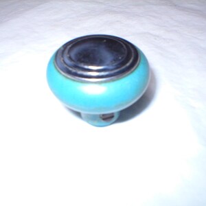 May include: A turquoise cabinet knob with a chrome-colored top and a silver-colored ring. The knob has a rounded shape and a small base with a screw. The knob is likely for a drawer or cabinet.