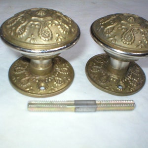 May include: Two antique brass door knobs with ornate floral designs and a silver rim. The knobs sit on matching circular bases. A threaded rod is positioned below the knobs, likely for installation. The hardware is likely vintage.