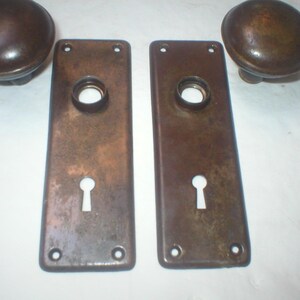 May include: A pair of dark brown, weathered metal door knobs with matching rectangular backplates. Each backplate features a central circular opening for the spindle and a lower keyhole cutout, with four small screw holes at the corners.