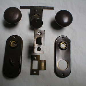 May include: A set of antique door hardware in a dark bronze finish. The set includes two round door knobs, two oval-shaped plates, a strike plate, a latch, and a keyhole cover.