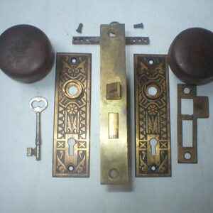May include: A brass door hardware set including two dark brown round knobs, two rectangular plates with intricate floral and geometric engravings, a central mortise lock mechanism, a skeleton key, a strike plate, and two small screws.