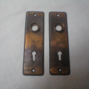 May include: Two antique bronze-toned door plates. Each rectangular plate features a keyhole, a circular opening, and two screw holes. The plates have a weathered appearance, suggesting age and use. Ideal for restoration projects or adding vintage charm.