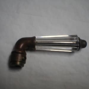 May include: A decorative, angled cabinet pull with a clear, ribbed glass handle and a bronze-colored metal base. The handle is rectangular with vertical ridges. The base has a curved elbow shape and a small, round end.