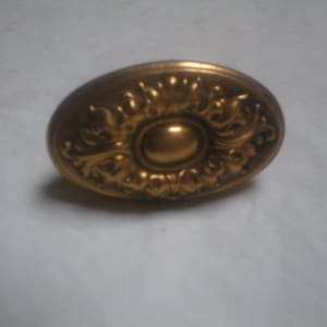 May include: Oval-shaped, antique brass door knob with ornate floral detailing. The knob features a central, raised oval surrounded by swirling, symmetrical patterns. A classic hardware piece.