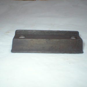 May include: A rectangular, cast iron object with a dark, aged appearance. It features two circular holes on the top surface and a raised, rectangular base. The object appears to be a vintage hardware component.