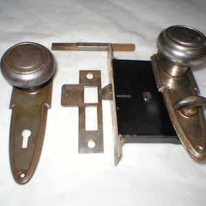 May include: A vintage door knob set with a silver finish and a keyhole. The set includes a knob, a strike plate, and a latch. The knob is round with a decorative design. The strike plate is rectangular with a keyhole. The latch is a small metal piece that fits into the strike plate.