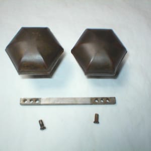 May include: Two antique bronze-colored cabinet knobs with a geometric design. A metal bar with multiple holes and two screws are also present. The knobs have a hexagonal shape and a slightly weathered appearance.