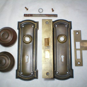 May include: A set of antique brass door hardware, including two knobs, two plates, a strike plate, and two screws. The hardware has a dark patina and a decorative design.