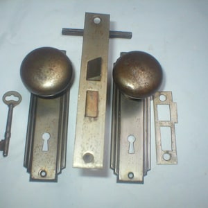 May include: A set of antique brass door hardware including two round knobs, two rectangular backplates with keyholes, a central mortise lock mechanism with a protruding latch, a separate strike plate, and a single skeleton key for door operation.
