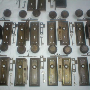 May include: An assortment of antique bronze door hardware, including doorknobs, faceplates, and keys. The hardware is arranged on a white surface, showcasing various styles and sizes. The collection appears to be from a vintage era, with signs of wear.