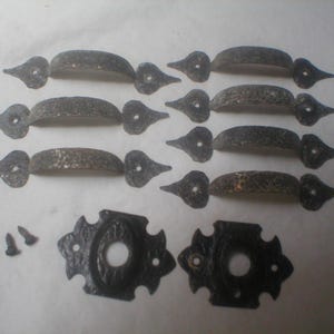 May include: A collection of antique-style, dark metal hardware pieces. The set includes several handles with decorative ends, matching backplates, and screws. The hardware has a textured finish, suggesting a rustic or vintage aesthetic, suitable for furniture or doors.