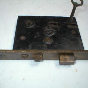 May include: Antique black metal door lock with a brass-colored strike plate. A skeleton key is inserted in the keyhole. The lock has a rectangular shape with visible screw heads and a weathered appearance, suggesting age.