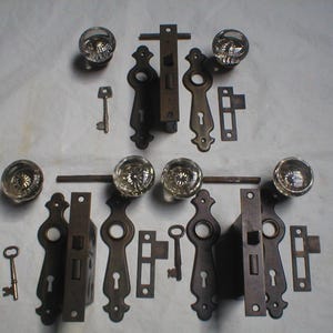 May include: An assortment of antique door hardware, including glass doorknobs, keys, and various metal latches and plates. The hardware has a vintage, aged appearance, with a mix of clear glass and dark metal finishes.