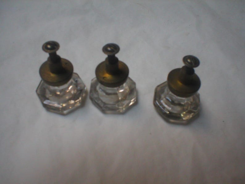 Puede incluir: Tres pomos de cristal transparente con forma octogonal y una base met&aacute;lica de color lat&oacute;n. Cada pomo cuenta con una peque&ntilde;a fijaci&oacute;n de tornillo en el centro, proporcionando un componente de herraje funcional para armarios o muebles.