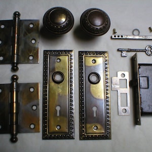 May include: A set of antique brass door hardware, including two hinges, two door knobs, two escutcheon plates, a key, a strike plate, and a door lock. The hardware is in good condition with some patina.