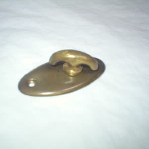 May include: A brass-colored, oval-shaped hook with a rounded handle. The hook is mounted on a matching oval base with a single hole for mounting. The hardware is likely for a door, cabinet, or other furniture.