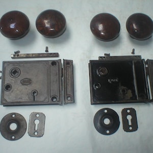 May include: Two sets of antique rim locks with dark brown round wooden knobs, rectangular metal lock bodies, matching skeleton keys, circular escutcheons, and rectangular keyhole covers. The hardware components are arranged in two distinct groups.