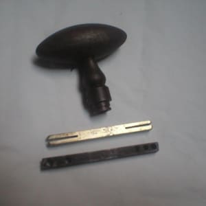 May include: A dark brown, oval-shaped metal door knob with a central stem and a cylindrical base. Below the knob are two separate metal components: a thin, flat, silver-toned rectangular bar with slotted ends and a dark, narrow rectangular metal strip.