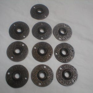 May include: A collection of ten antique bronze-colored door rosettes. Each rosette features an ornate, embossed floral design around a central hole, with three screw holes. The rosettes are arranged on a white surface.