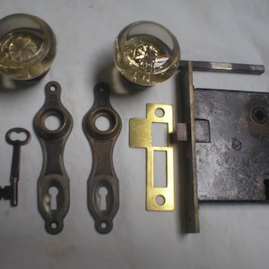 May include: A set of door hardware including two clear glass knobs with internal star patterns, two brass-toned metal escutcheon plates, a skeleton key, a brass strike plate, and a rectangular iron mortise lock mechanism with a square spindle.