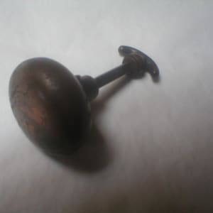 May include: A vintage bronze-toned door knob with an oval handle and a slender stem. The knob has a slightly textured surface and a small, curved handle on the opposite end of the stem. The hardware is likely for a cabinet or door.