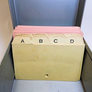 Puede incluir: Una caja metálica de archivo gris abierta que contiene una pila de tarjetas de índice rosas y un juego de tarjetas de índice amarillas con pestañas etiquetadas A, B, C y D. La caja tiene un diseño sencillo.