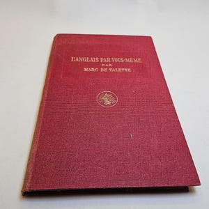 Peut inclure: Un livre rouge vintage intitulé "L'Anglais Par Vous-Même" par Marc de Valette. La couverture présente des lettres dorées et un emblème circulaire. Le livre a une surface texturée et montre des signes d'âge.