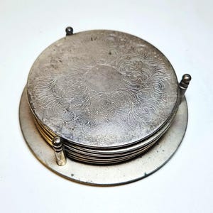 May include: A stack of circular silver-toned metal coasters held in a matching metal holder. The top coaster features an intricate, engraved floral scrollwork pattern. The holder has three vertical posts with small spherical finials at the top.